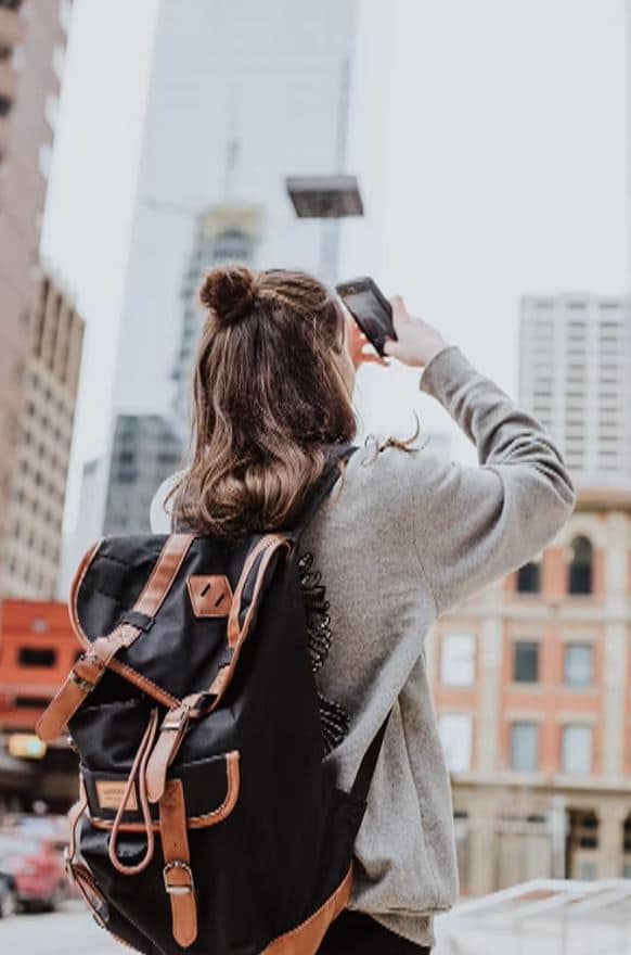 Mulher de costas com uma mochila e o celular nas mãos tirando foto de uma paisagem na cidade.