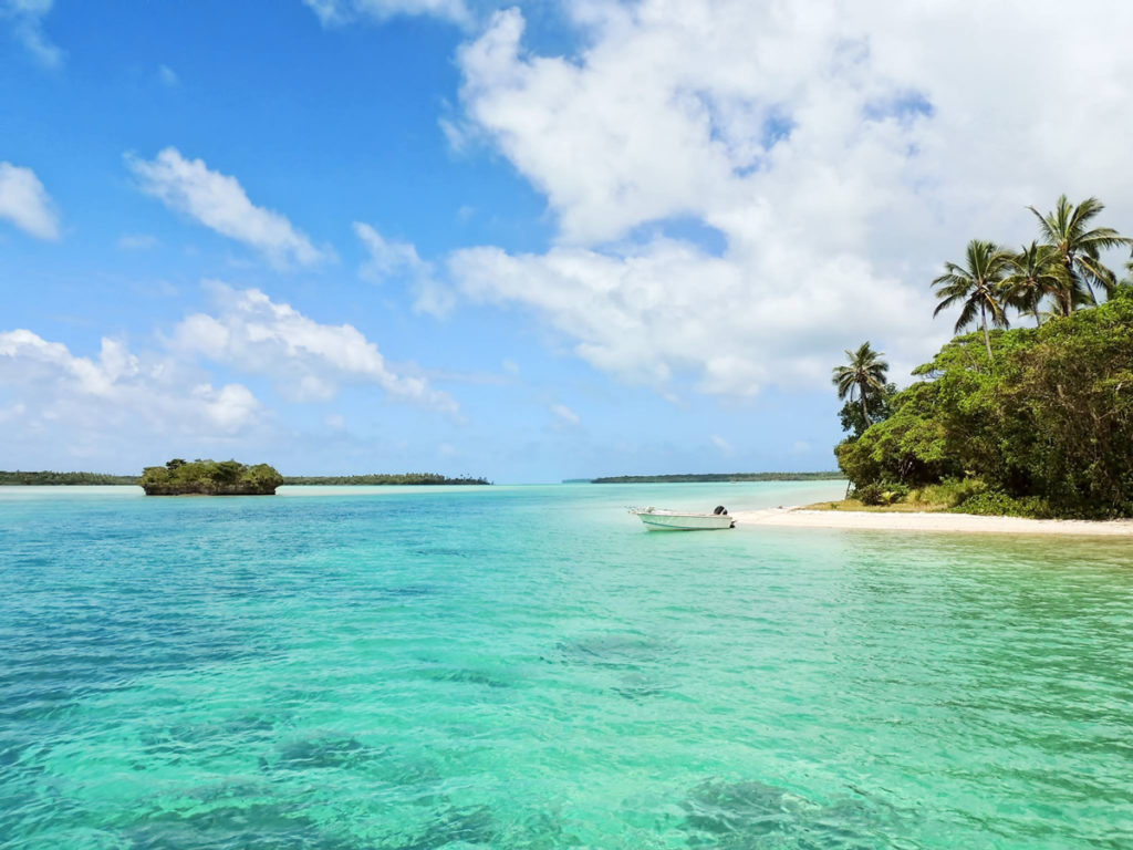 Foto da categoria "praias": uma praia com mar azulado, pequena faixa de areia ao lado direito e um pequeno barco ancorado.